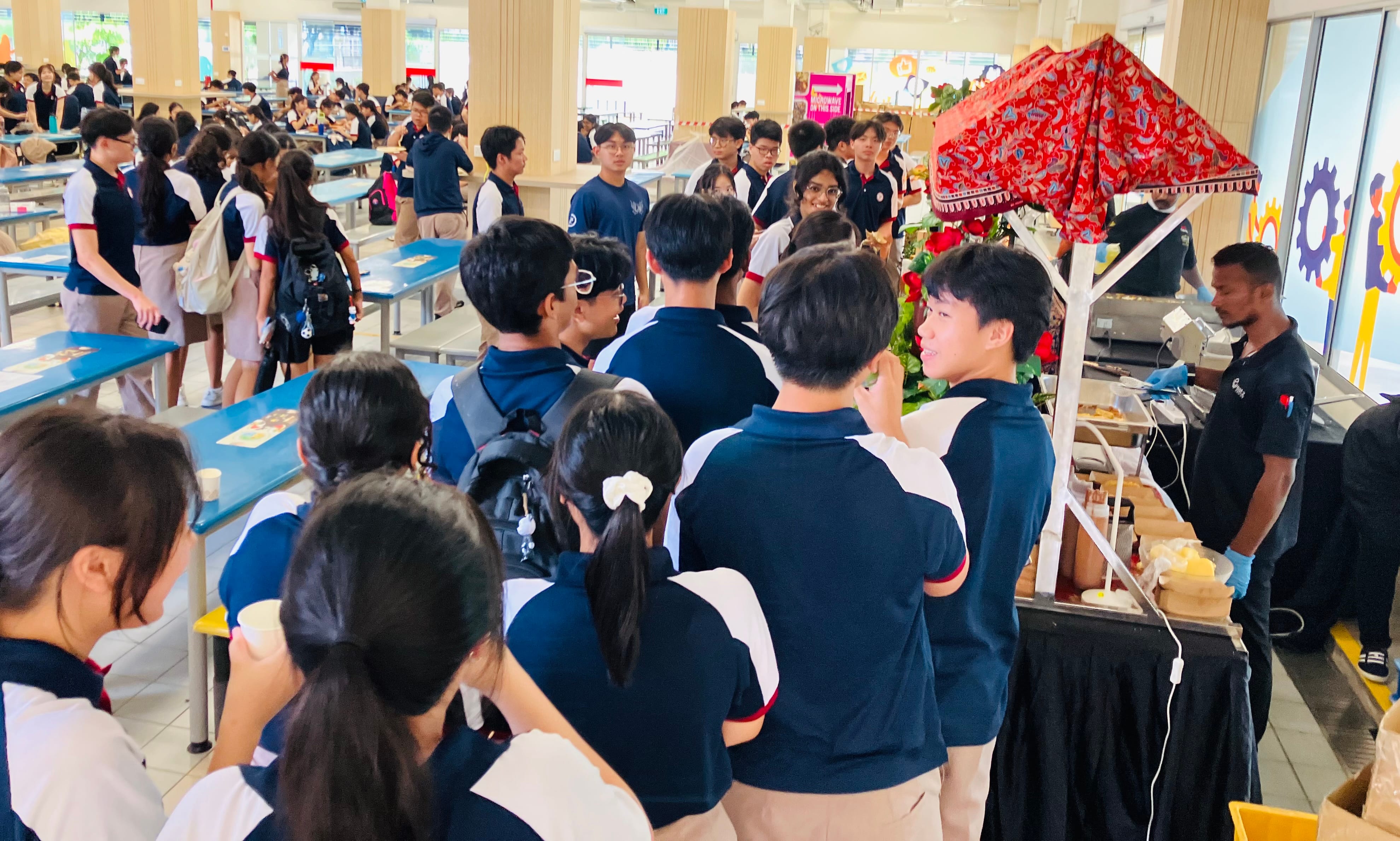A large crowd of students in school uniforms queue eagerly at a food booth decorated with a colourful red batik-patterned canopy in a school canteen. A staff member wearing gloves serves food at the stall while students wait in a long line stretching across the busy canteen.